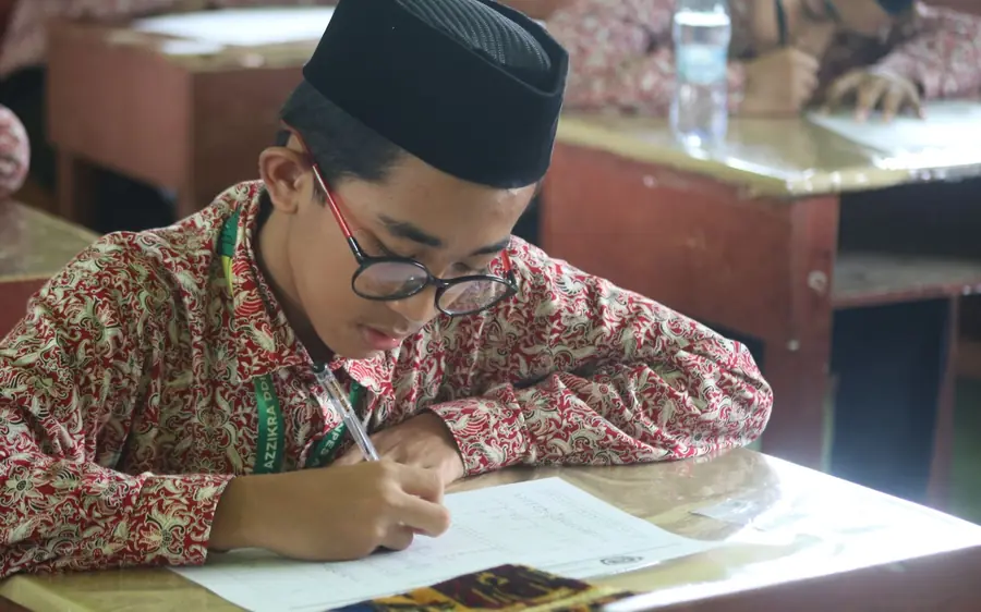 Seorang murid lelaki berkopiah menulis latihan di meja